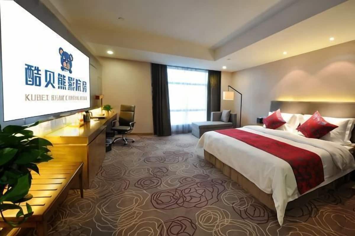 A hotel room with a bed, desk, chair, and a large screen displaying hotel branding.