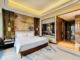 Una habitación de hotel moderna con una cama grande, una mesita de noche, un espejo montado en la pared y un baño visible a través de una puerta abierta.