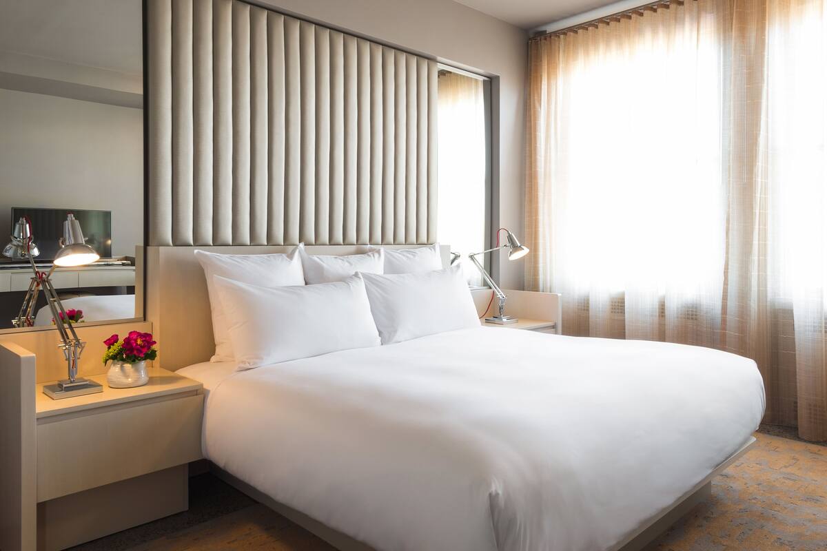 A neatly made bed with white linens, a bedside table with a lamp, and a vase with flowers.