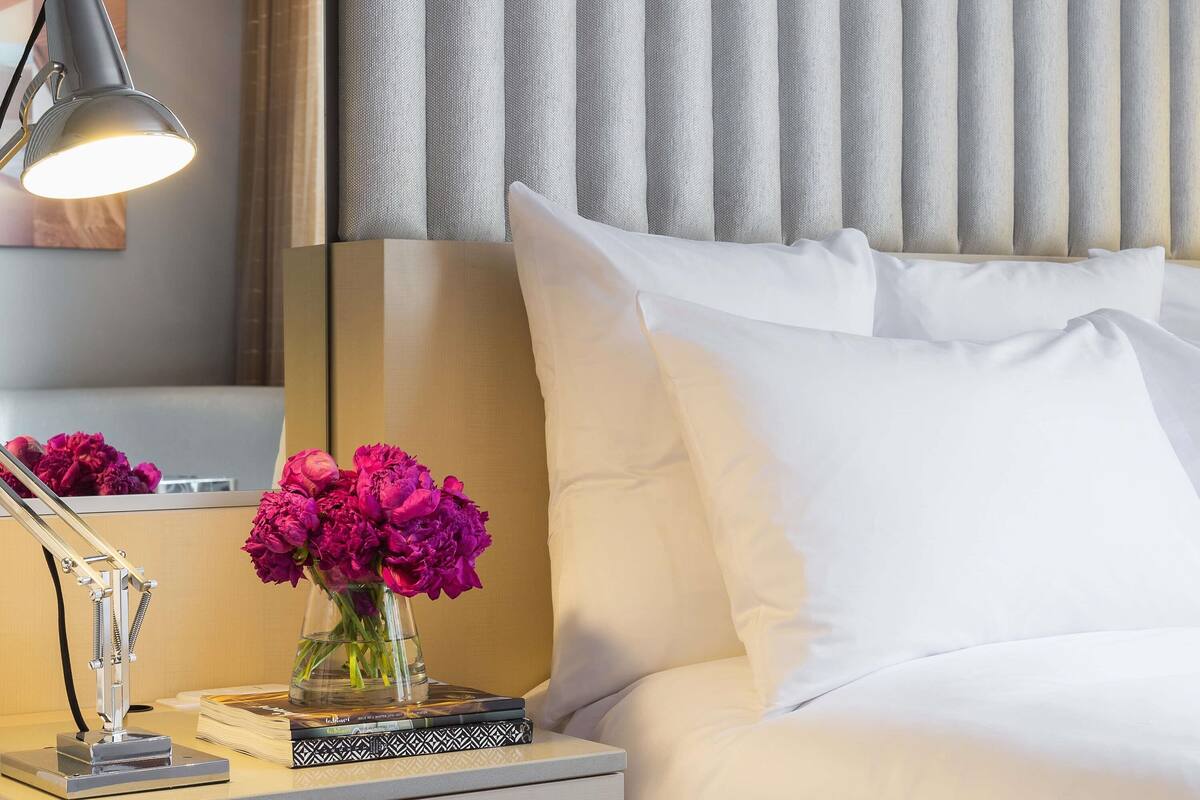A hotel room with a bed, a nightstand with a lamp, a vase of flowers, and a window with curtains.