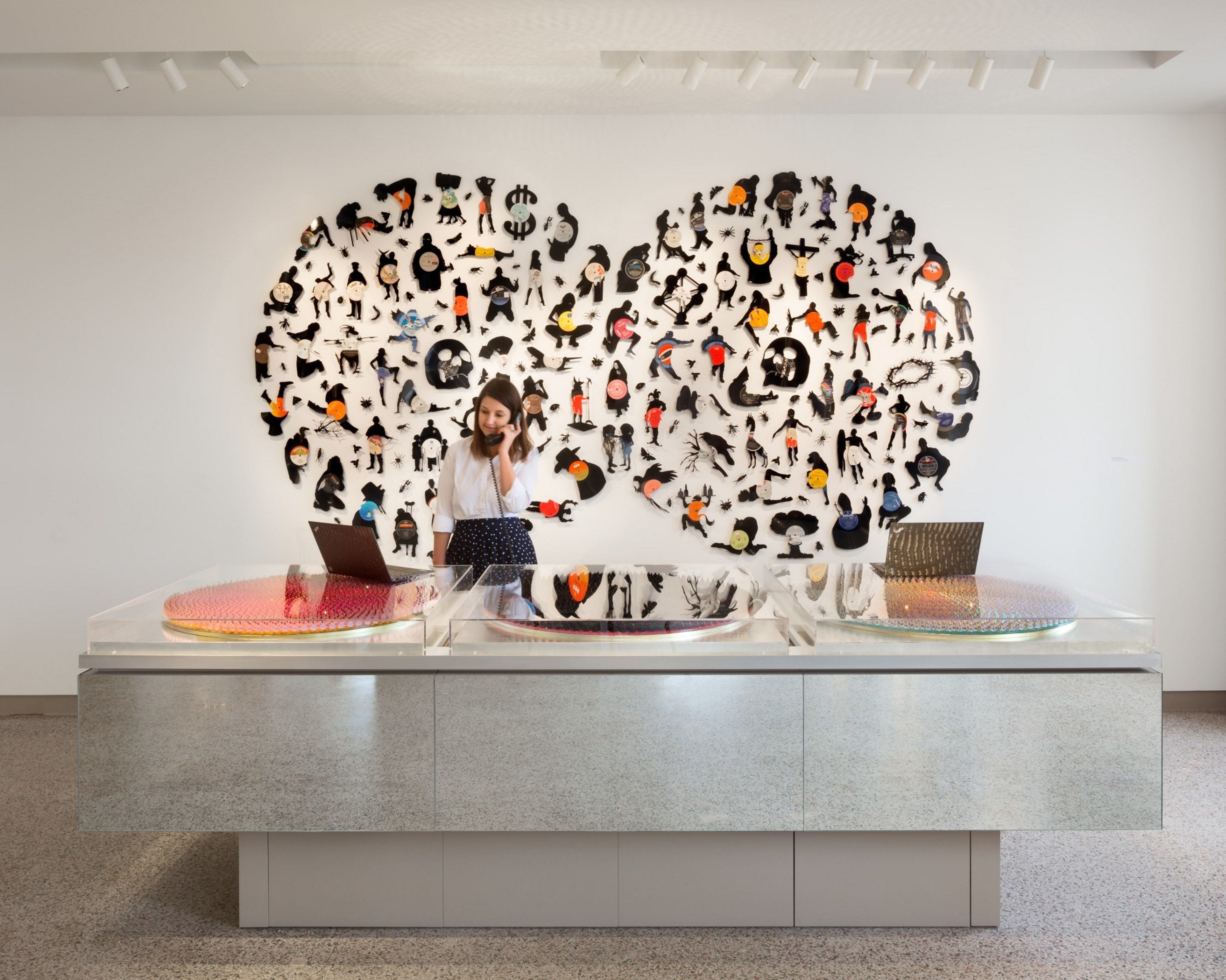 Un intérieur moderne avec une table centrale servant de présentoir, où sont disposés un ordinateur portable et un sac à main, le tout entouré d’un mur orné de nombreuses silhouettes découpées aux couleurs et aux formes variées.