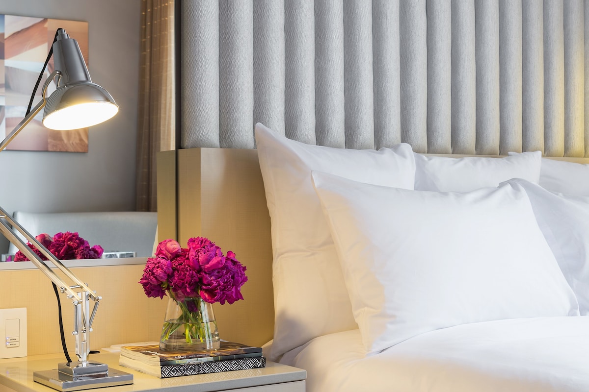 A neatly made hotel room with a bed, white pillows, a bedside table with a lamp, a vase of flowers, and a framed picture on the wall.