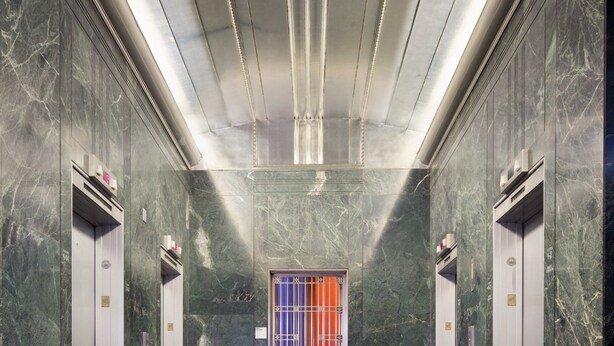 Un moderno vestíbulo de ascensores con paredes de mármol, un banco y un ascensor central con puerta colorida.