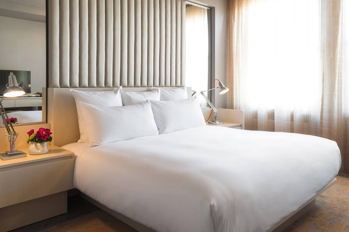 A neatly made bed with white linens, a bedside lamp, and a vase with flowers on the nightstand.