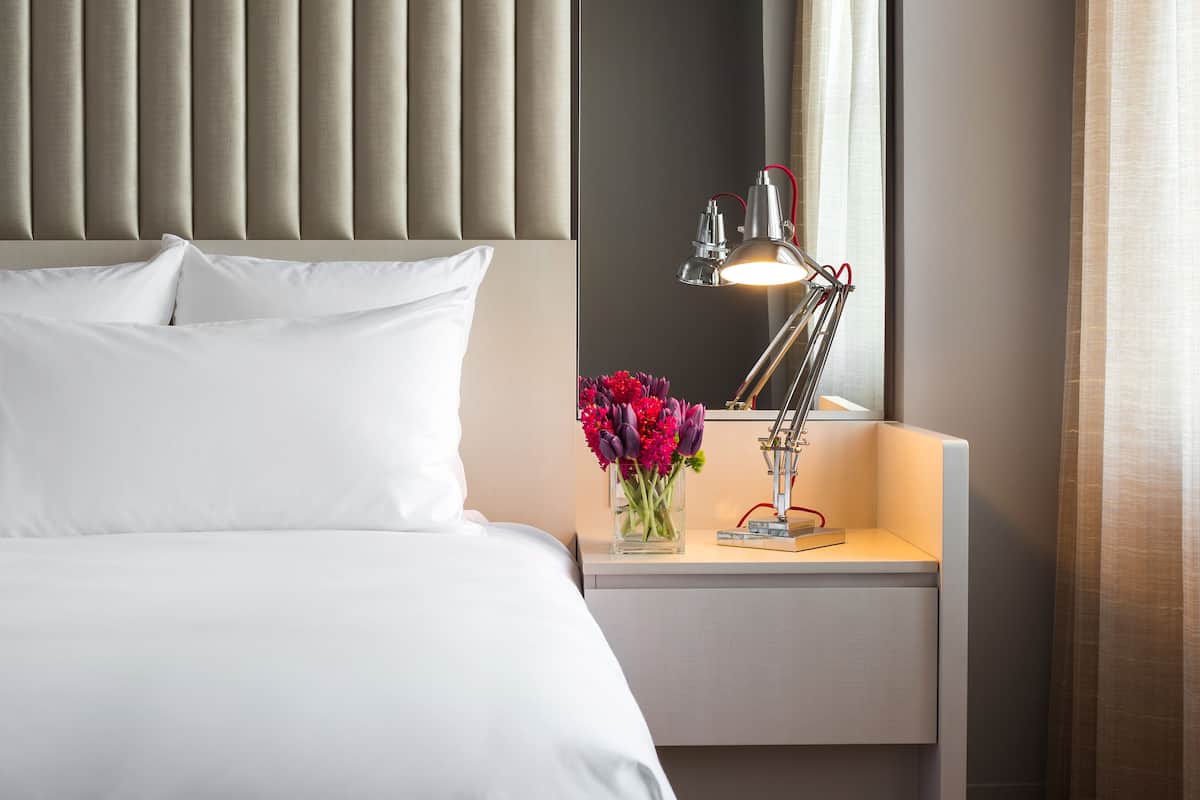 A neatly made bed with white linens, a bedside table with a lamp, and a vase of flowers.