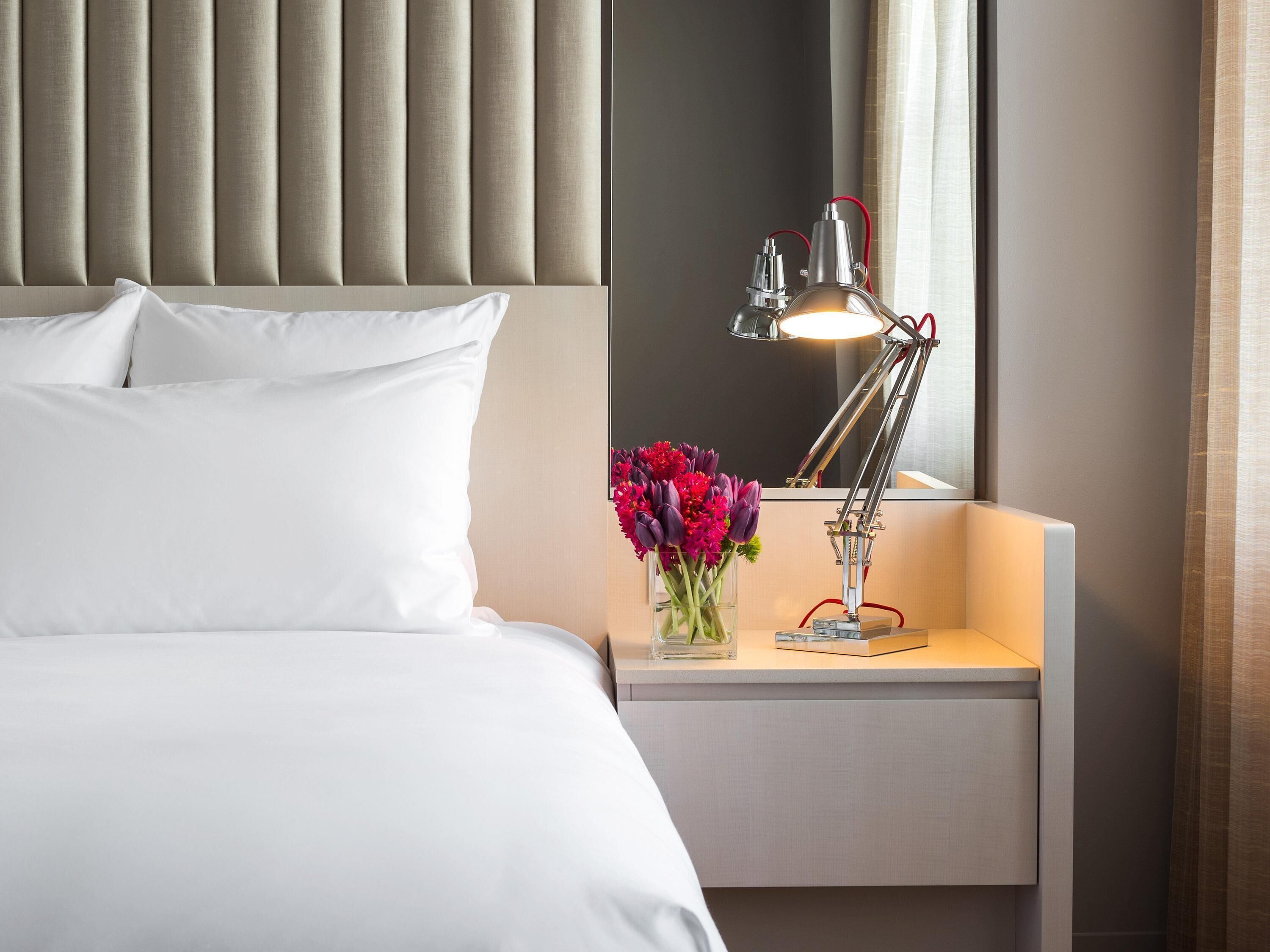 A neatly made bed with white linens, a bedside table with a lamp and a vase of flowers, and a headboard with vertical panels.