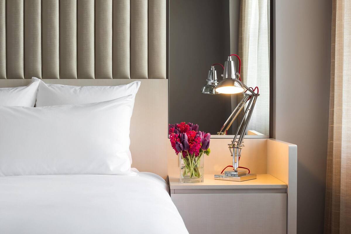 A neatly made bed with white linens, a bedside table with a lamp and a vase of flowers, and a headboard with vertical panels.