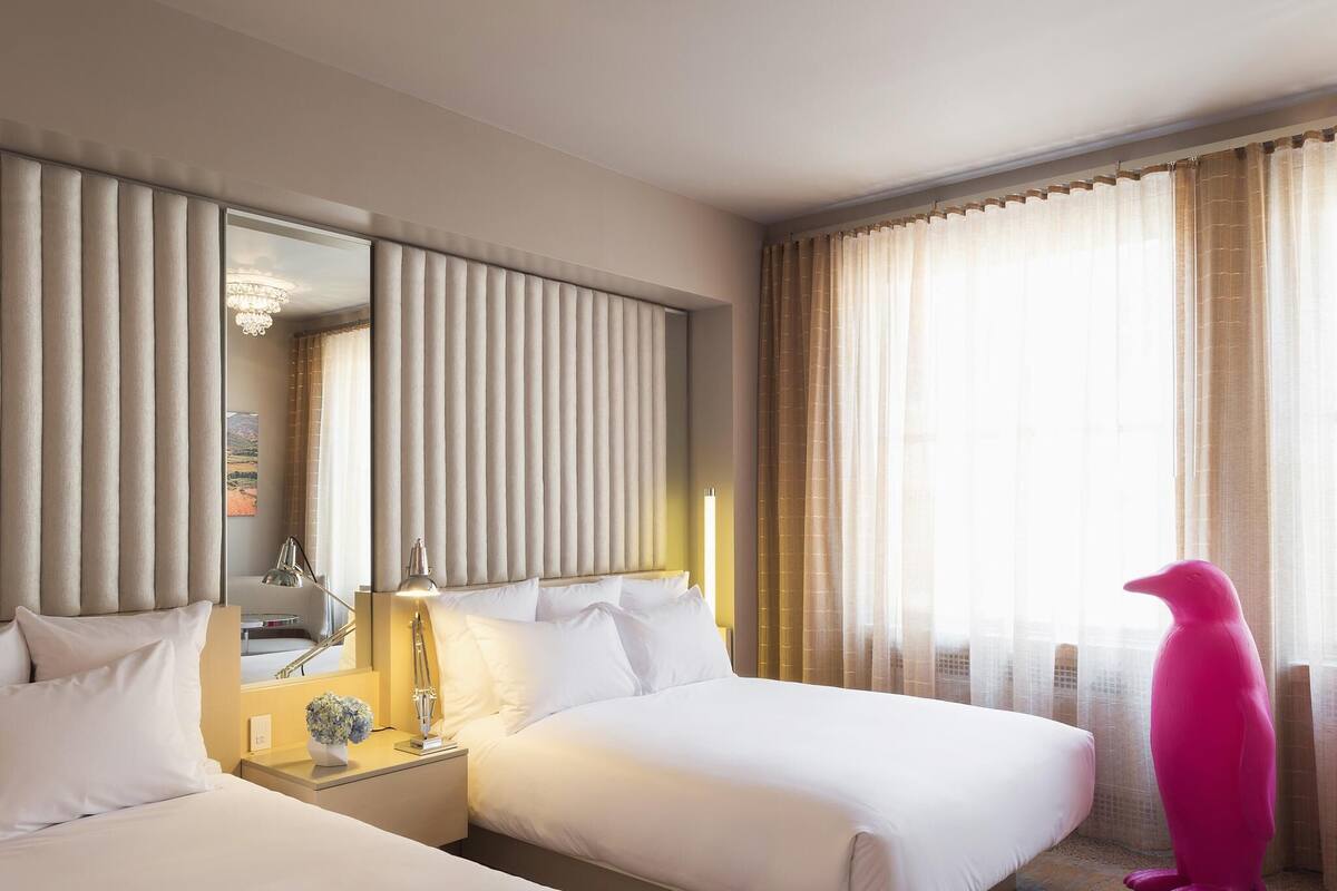 A hotel room with two beds, a nightstand, a lamp, a mirror, and a window with sheer curtains.