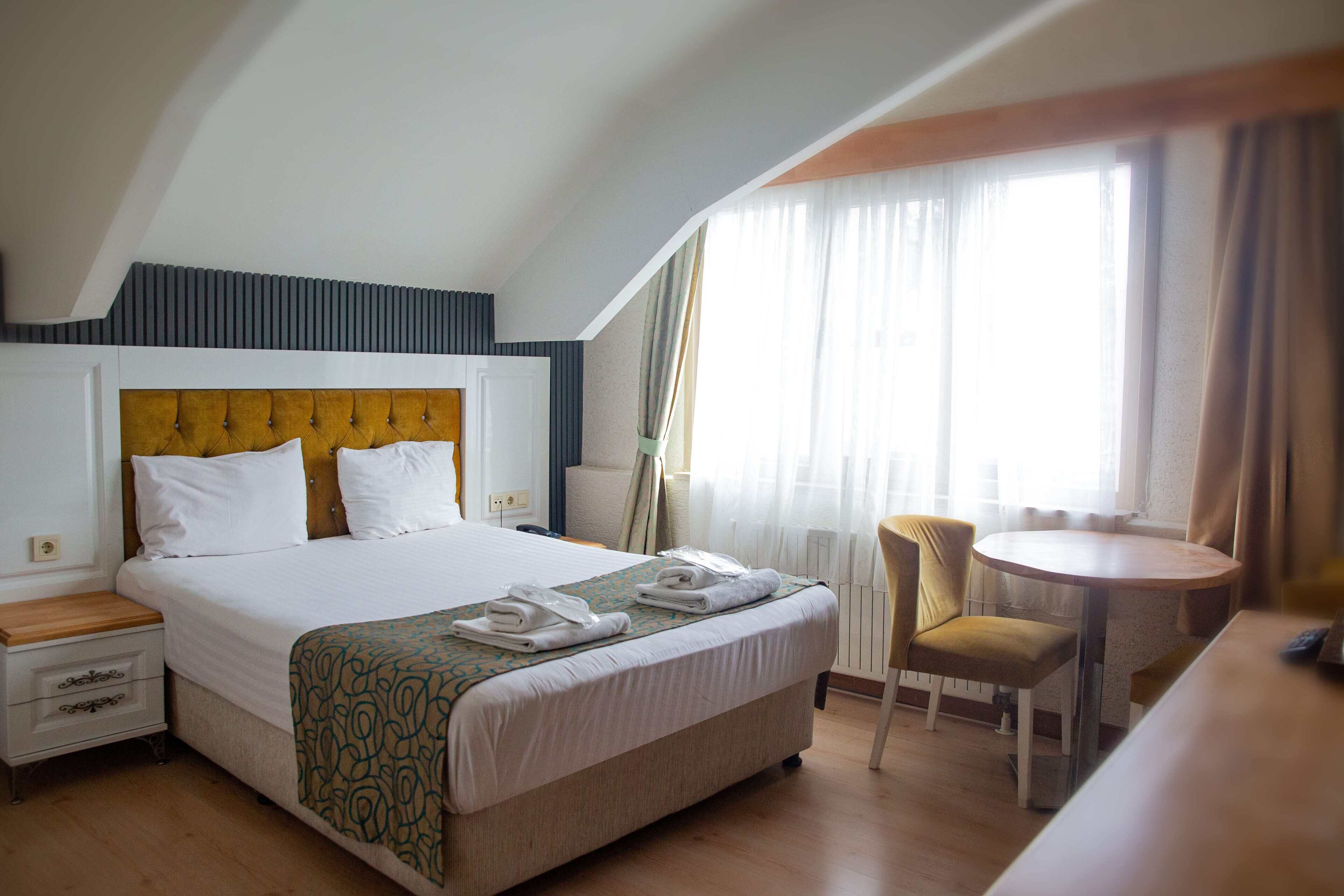 Habitación de hotel con una cama grande, una mesa redonda, una silla y un ventanal con cortinas.