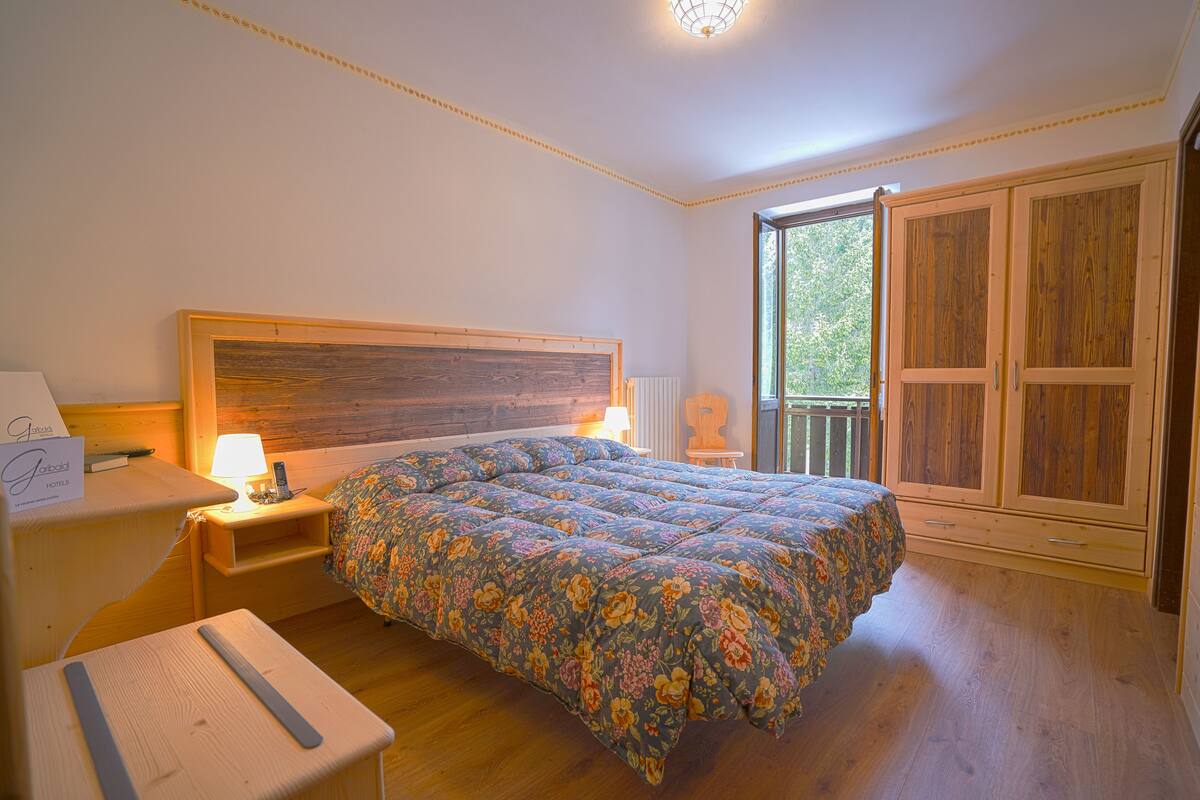 Ein Schlafzimmer mit einem hölzernen Kopfende, einer blumigen Tagesdecke, einem Nachttisch mit Lampe und Blick ins Freie durch ein Fenster.