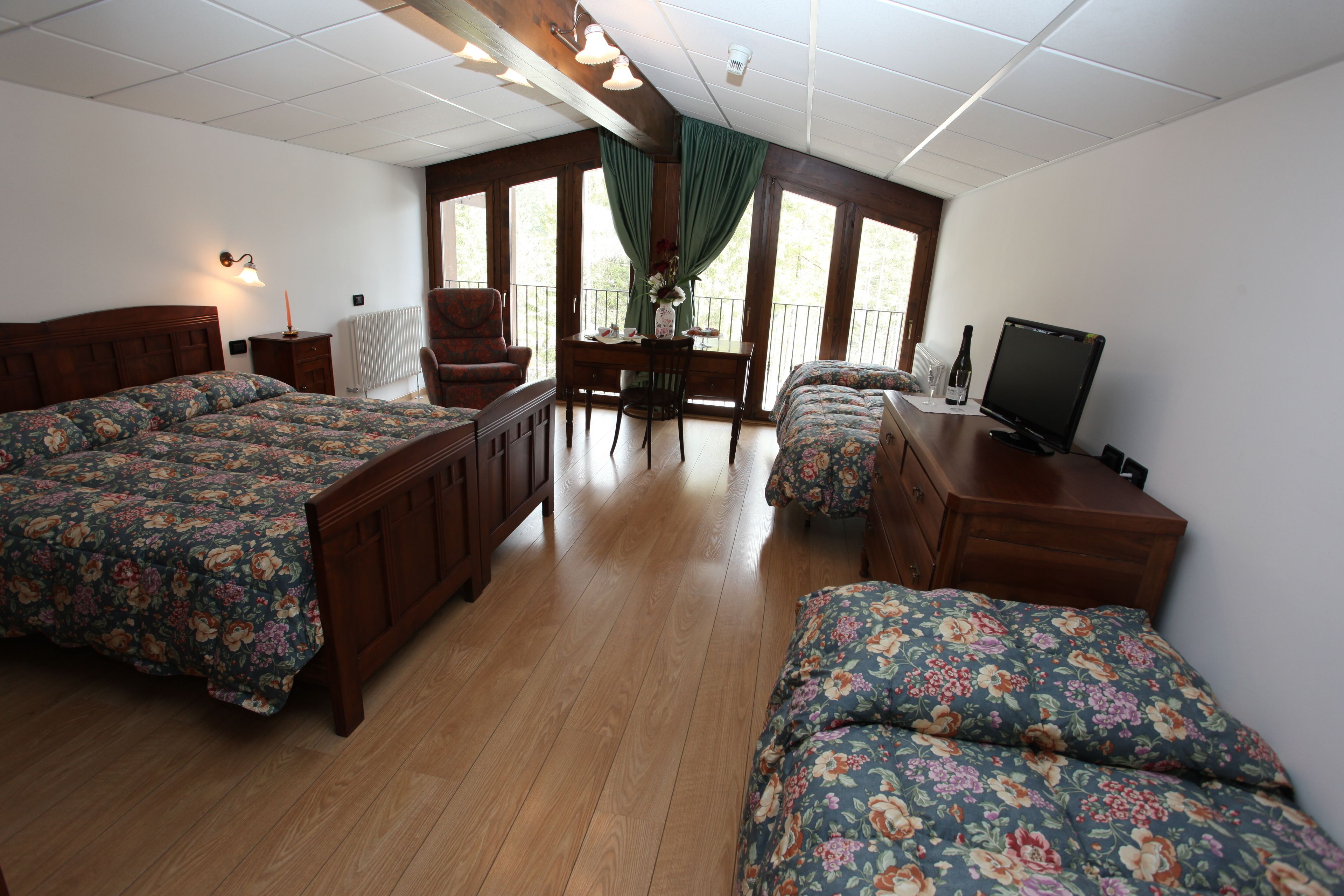 A bedroom with two beds, a wooden desk, a chair, a TV, and large windows with curtains.