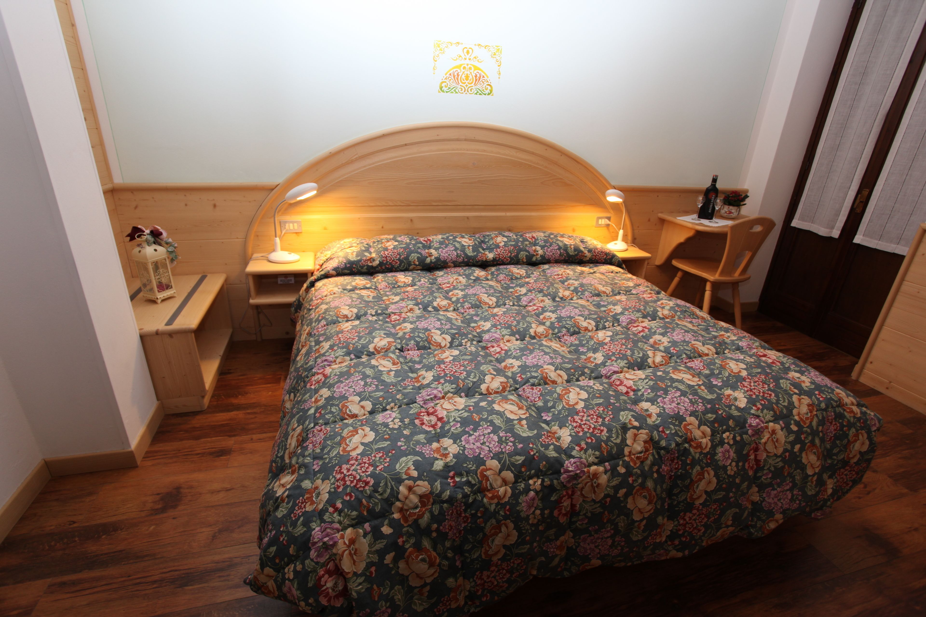 A bedroom with a floral bedspread, wooden headboard, bedside tables with lamps, a small table with a bottle, and a chair.