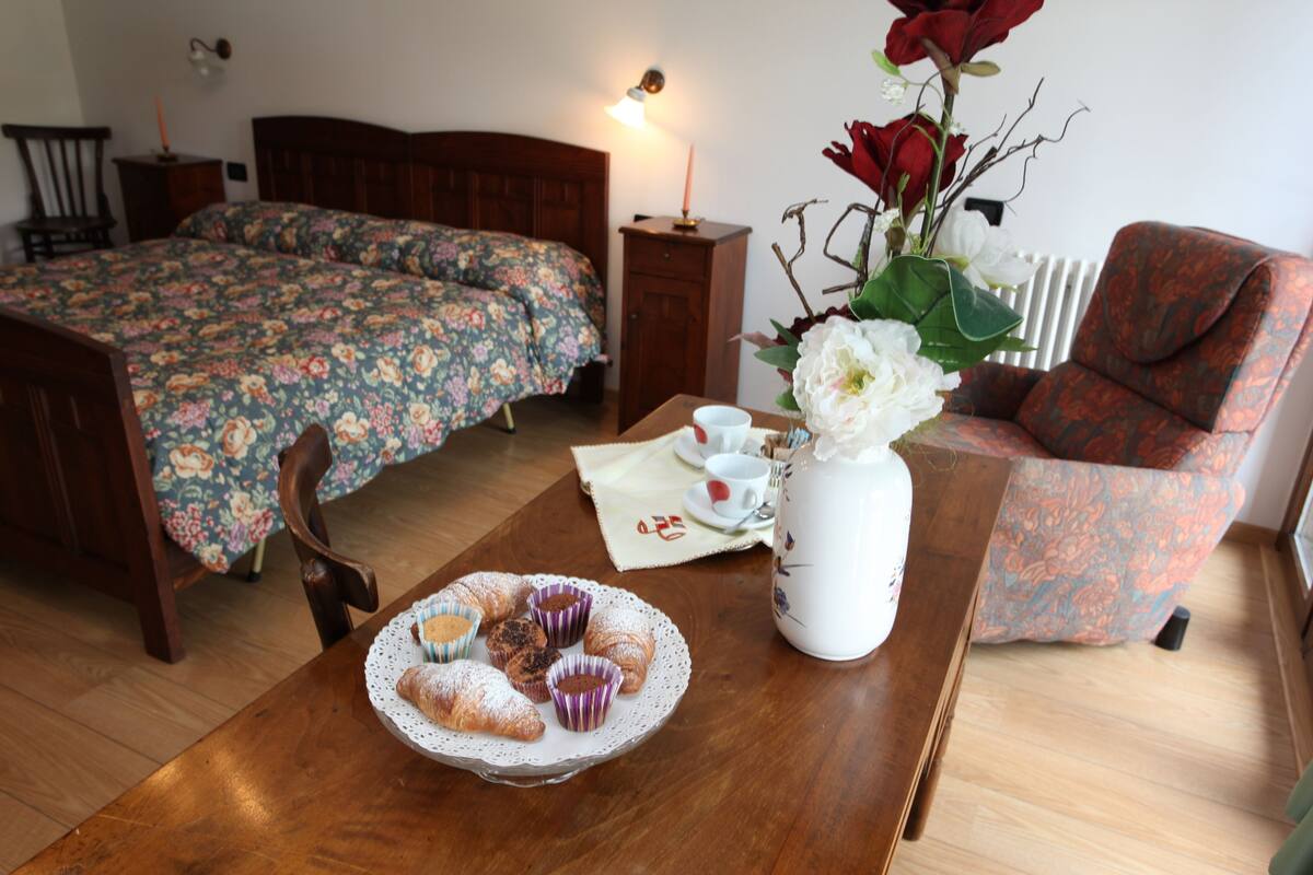 Ein Schlafzimmer mit einem Bett, einem Sessel, einem Tisch mit Gebäck, einer Blumenvase und einer Lampe.