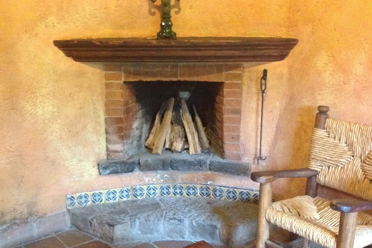 Une cheminée avec un manteau en briques et un chandelier sur le dessus. Devant la cheminée, on trouve une chaise en osier et une table en bois.