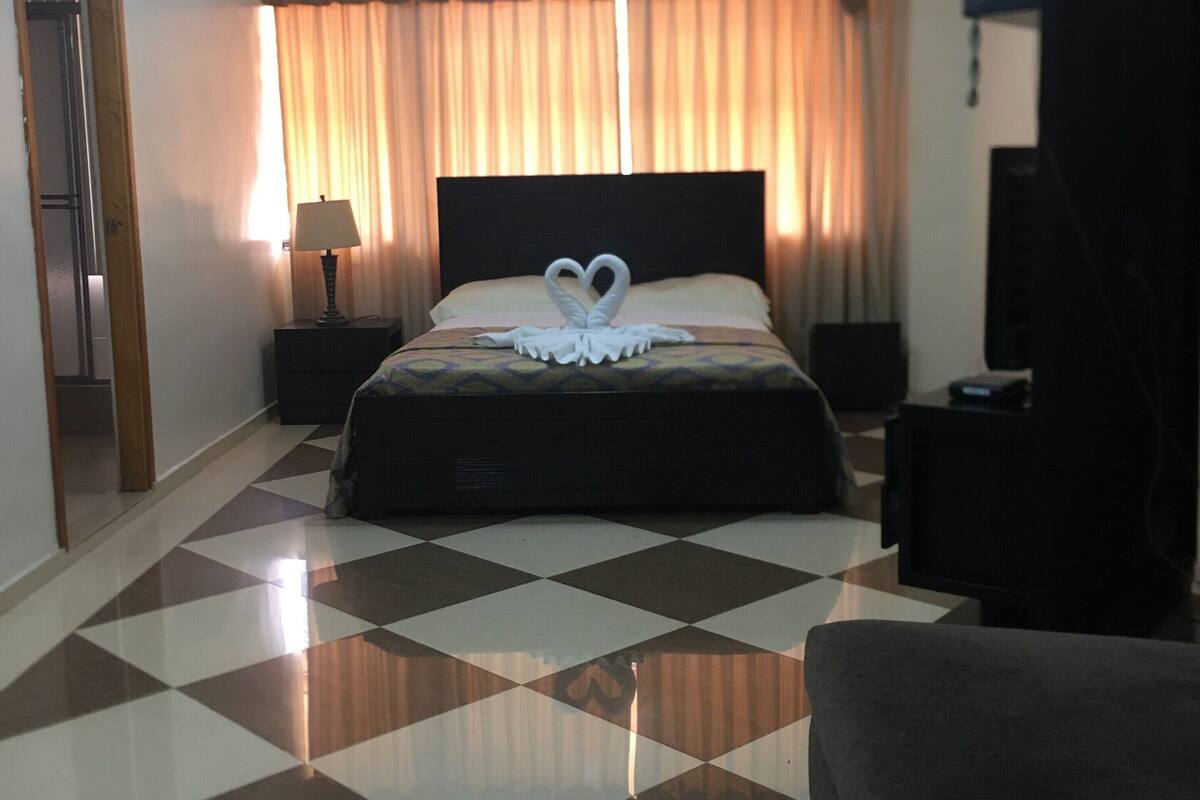 A hotel room with a checkered floor, a bed with a heart-shaped towel, a ceiling fan, and a television.