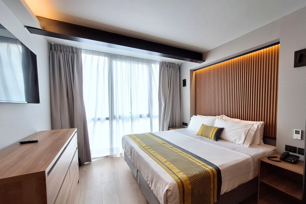 Quarto de hotel moderno com uma cama grande, uma escrivaninha de madeira, mesa de cabeceira com telefone e uma janela ampla com cortinas.