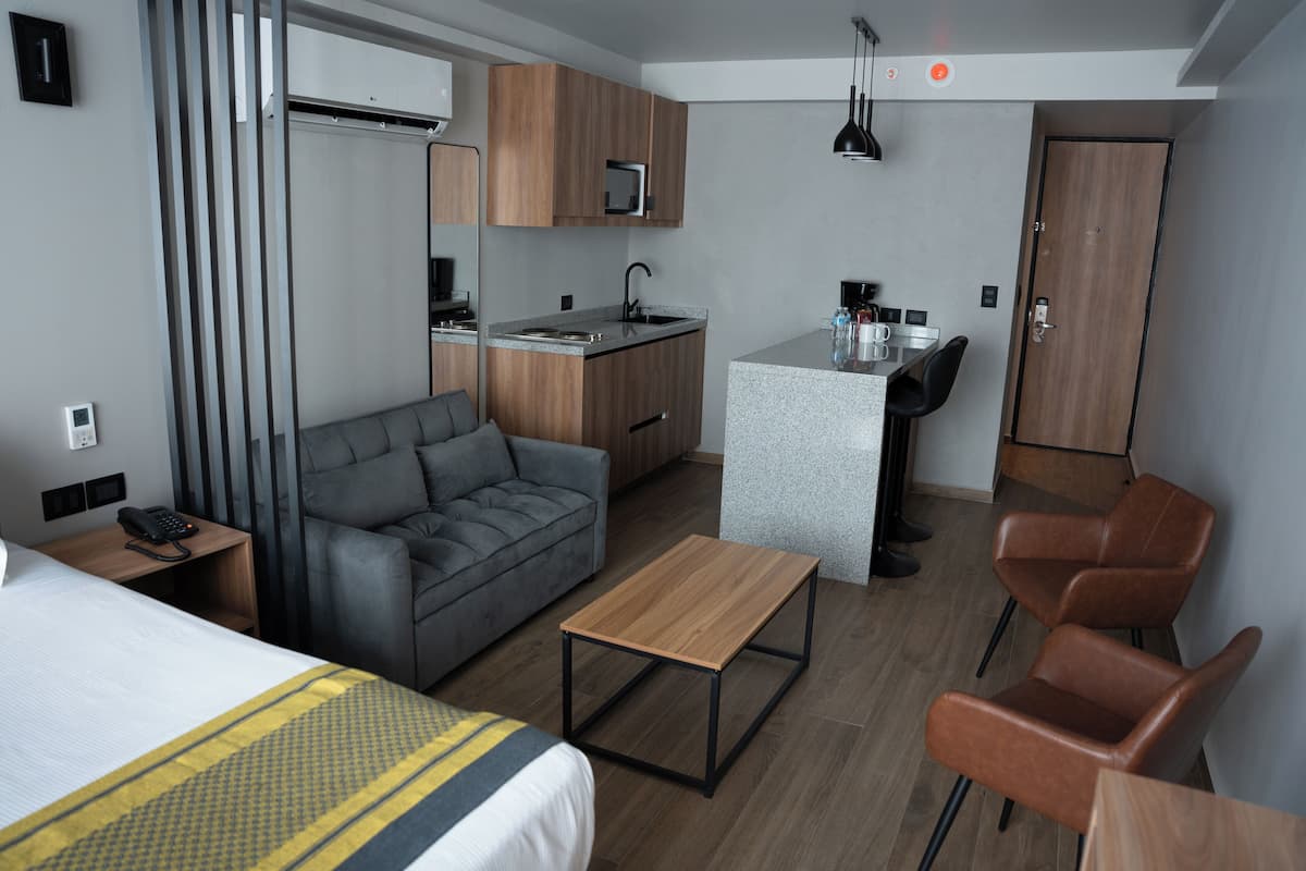 A compact hotel room with a bed, a sofa, a small kitchen area, and a seating area with chairs.