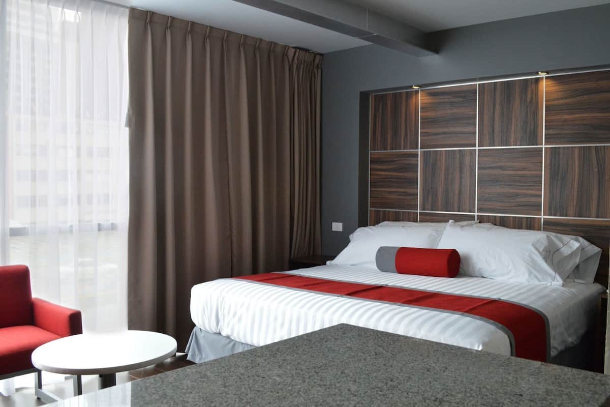 A hotel room with a bed, a red chair, a small round table, and a view of the city through sheer curtains.