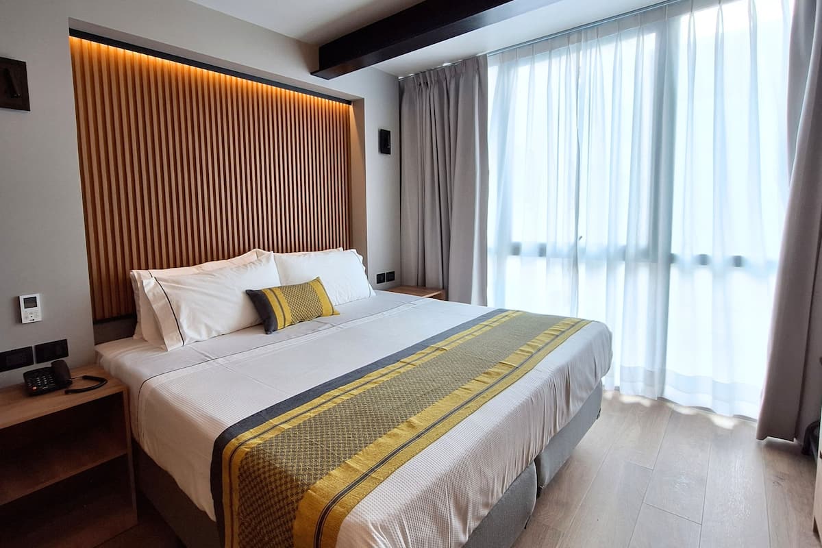 A hotel room with a large bed, a wooden headboard, a bedside table with a phone, and a window with sheer curtains.