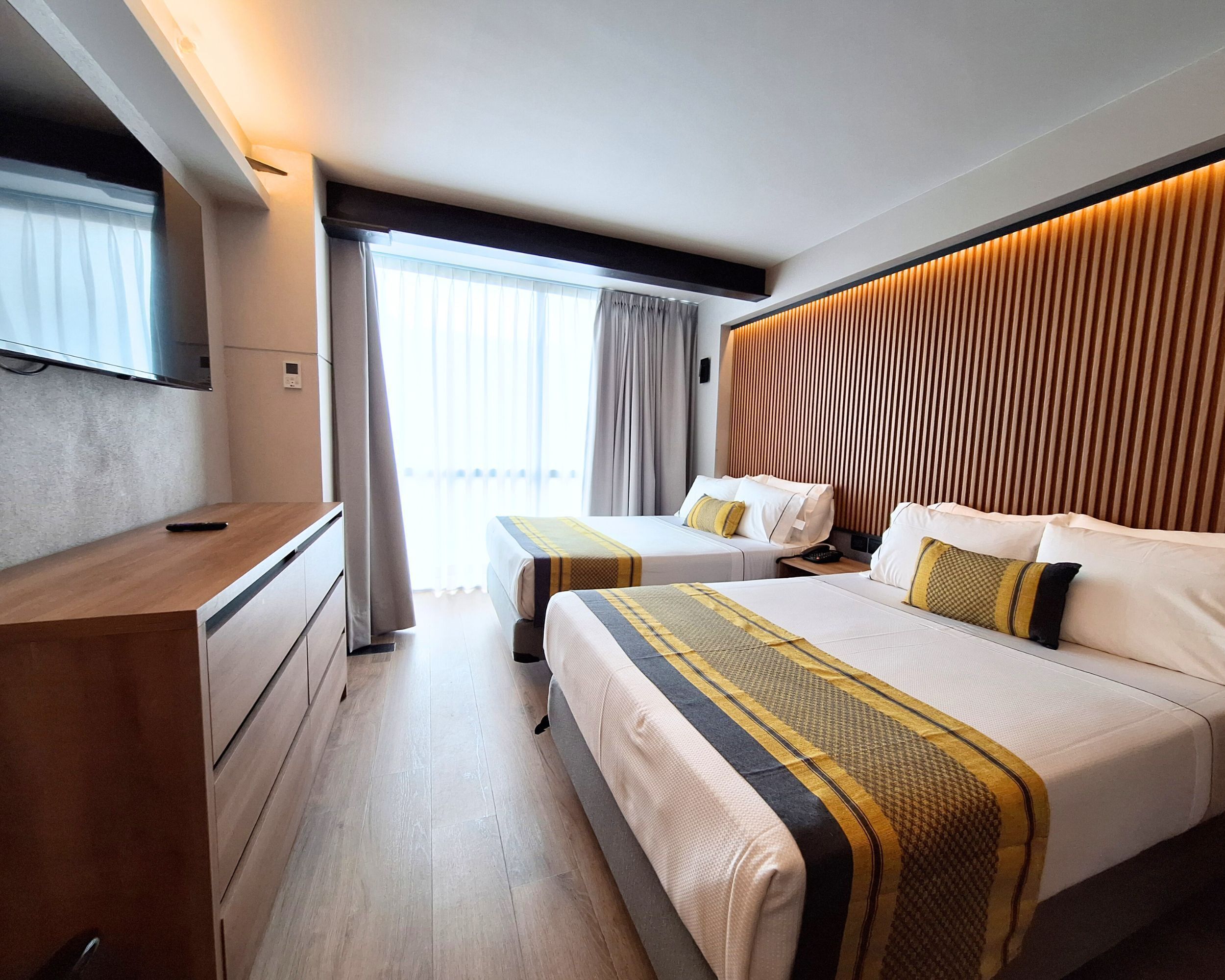 A modern hotel room with a wooden desk, two beds with matching bedding, a large window with curtains, and a wall-mounted TV.