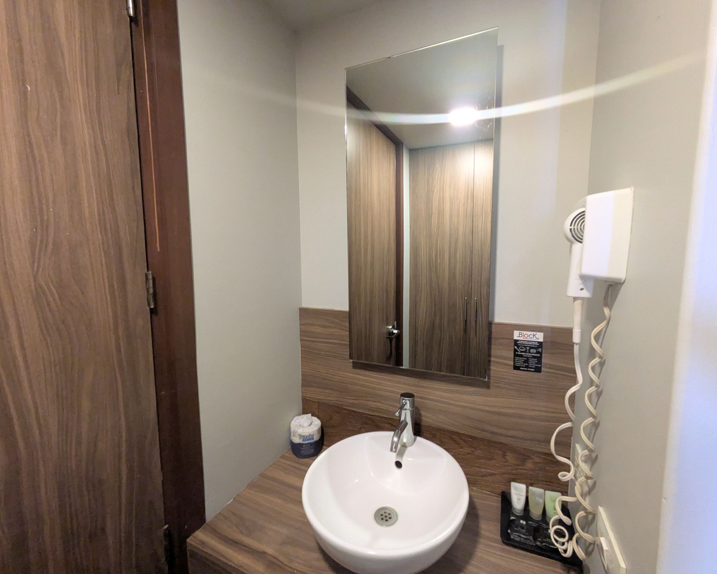 Une salle de bain avec un meuble-lavabo en bois, un lavabo blanc, un miroir et un sèche-cheveux fixé au mur.