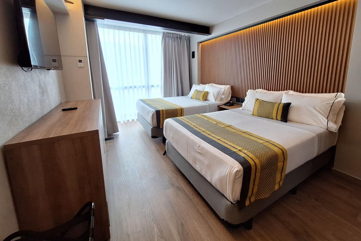 Quarto de hotel com duas camas, uma televisão fixada na parede, uma escrivaninha de madeira e uma janela grande com cortinas.