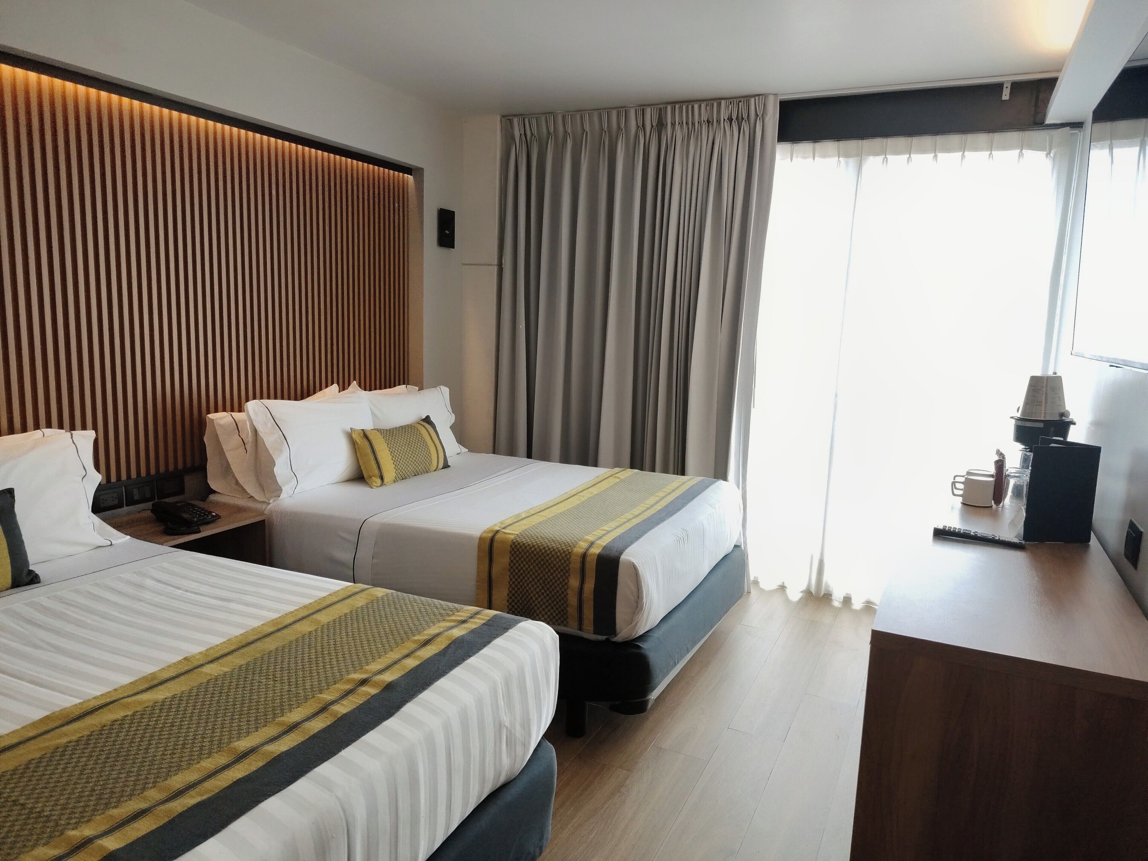 Habitación de hotel con dos camas, cabecero de madera, escritorio con lámpara y ventana con cortinas.