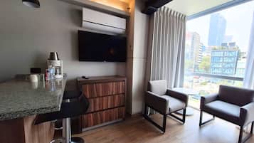 Una habitación de hotel moderna con área de cocina, televisor de pantalla plana, sofá y un amplio ventanal con vista a la ciudad.