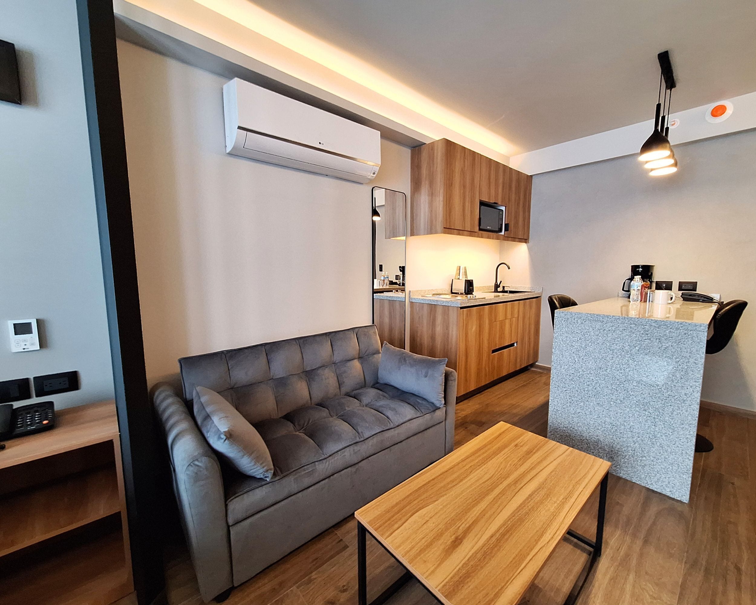 A compact living space with a sofa, a small wooden table, a kitchenette with a microwave, and a ceiling-mounted air conditioner.