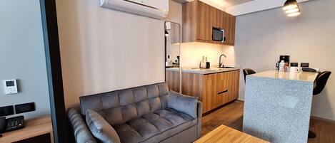Un espace de vie compact comprenant un canapé, une petite table en bois, une kitchenette équipée d’un four à micro-ondes et un climatiseur fixé au plafond.