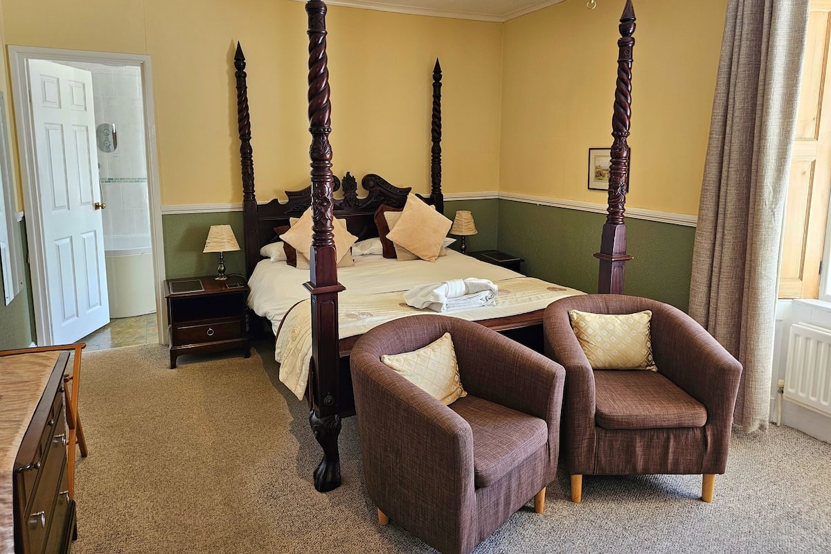 Uma cama com dossel e cabeceira de madeira, duas poltronas marrons, um criado-mudo com abajur e uma cômoda.