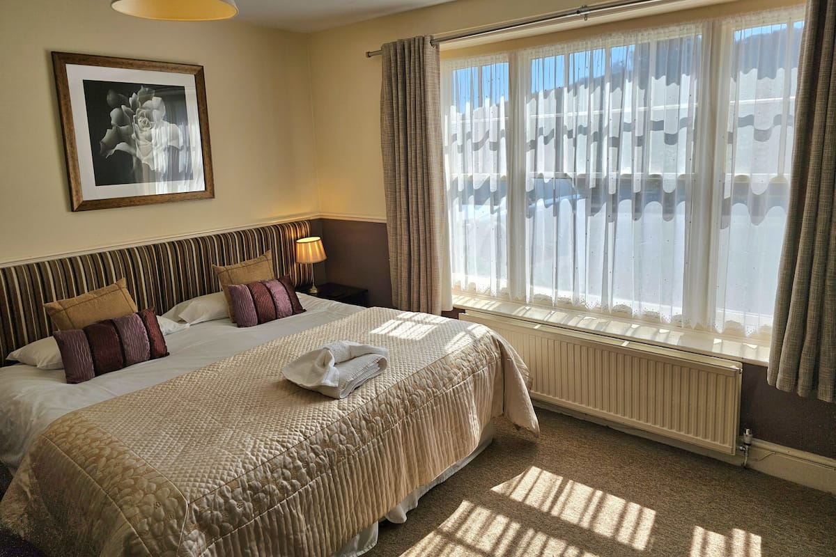 Quarto de hotel com uma cama grande, mesa de cabeceira com abajur, quadro na parede e janela com cortinas.