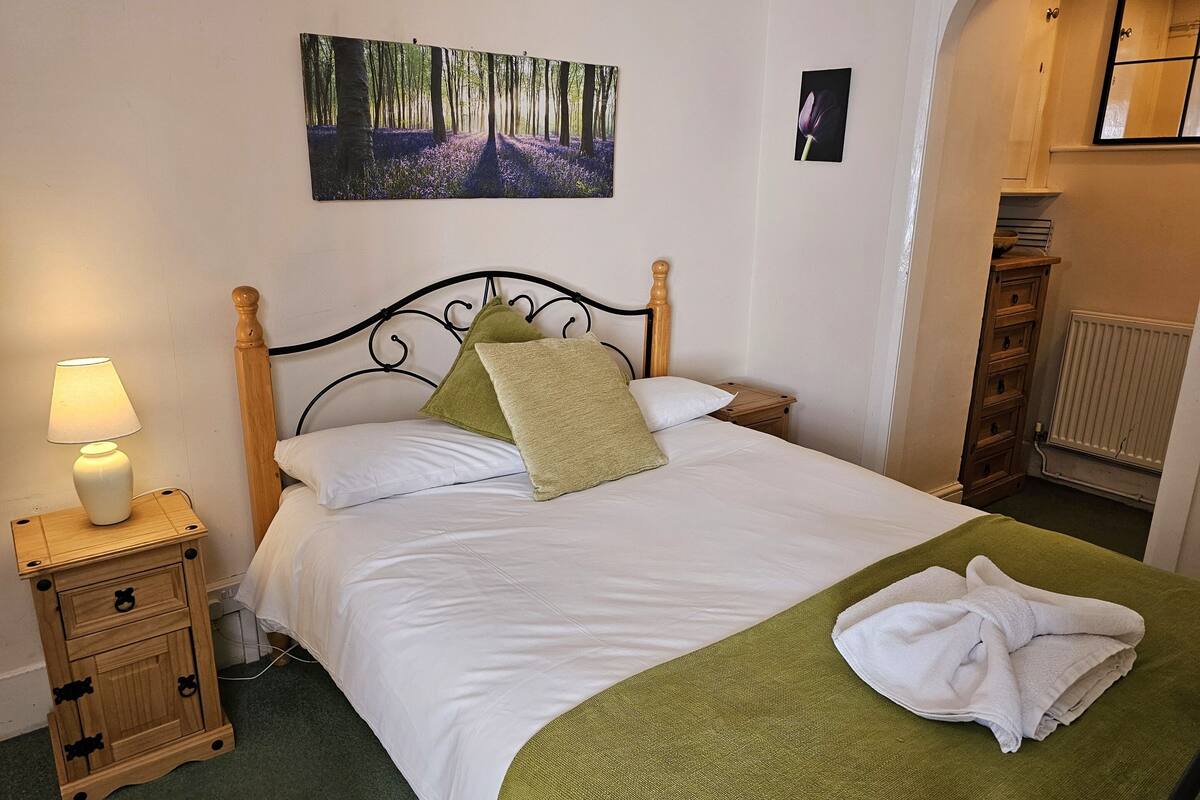 Cama bem arrumada com roupa de cama branca e manta verde, uma luminária de cabeceira e uma pequena mesa de cabeceira de madeira com gavetas.