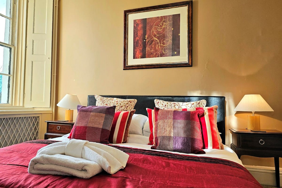 Cama bem arrumada com colcha vermelha e toalhas brancas, duas mesas de cabeceira com abajures, uma janela com vista para árvores e uma obra de arte emoldurada na parede.