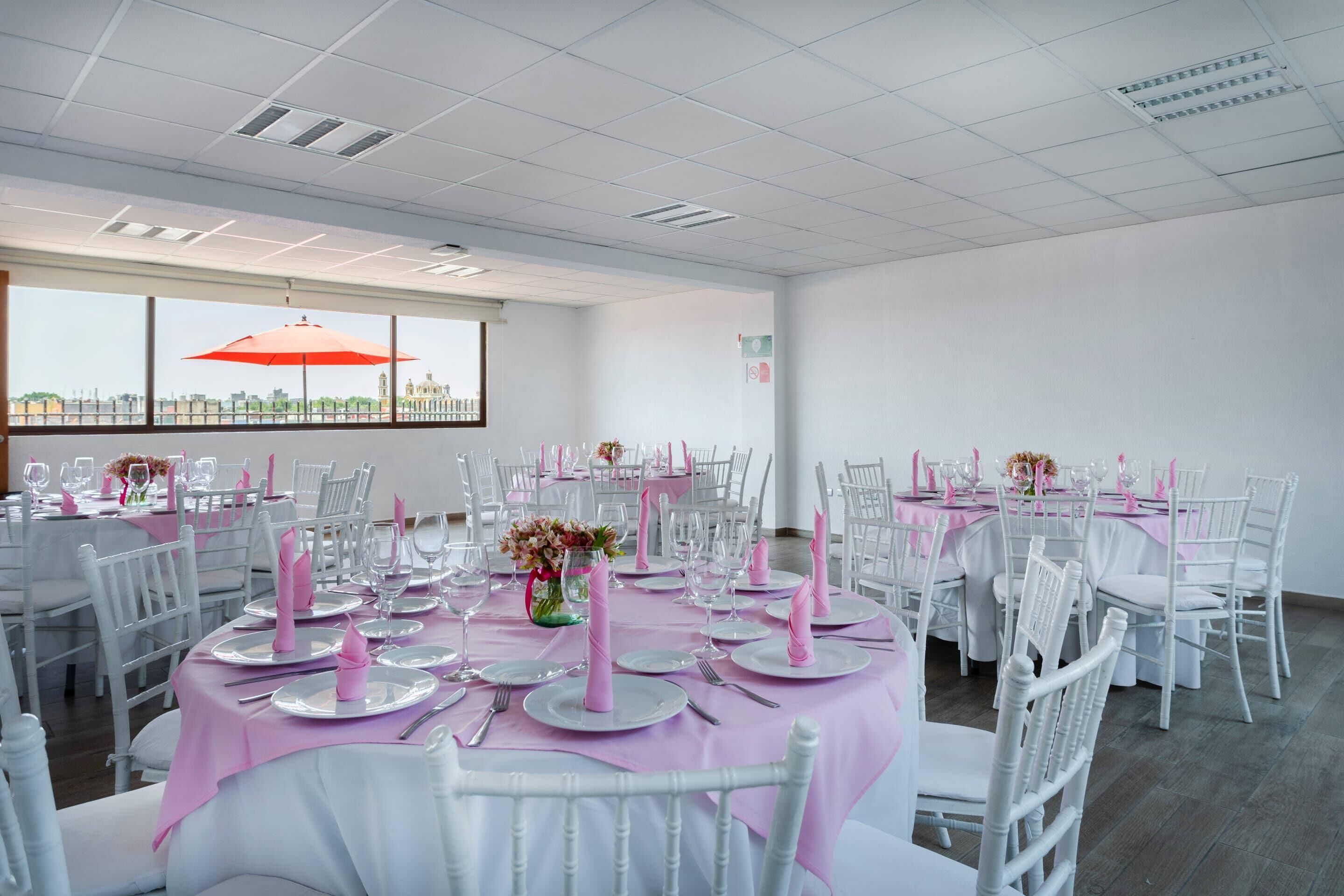 Un salón de banquetes con mesas redondas, sillas blancas y manteles rosas.