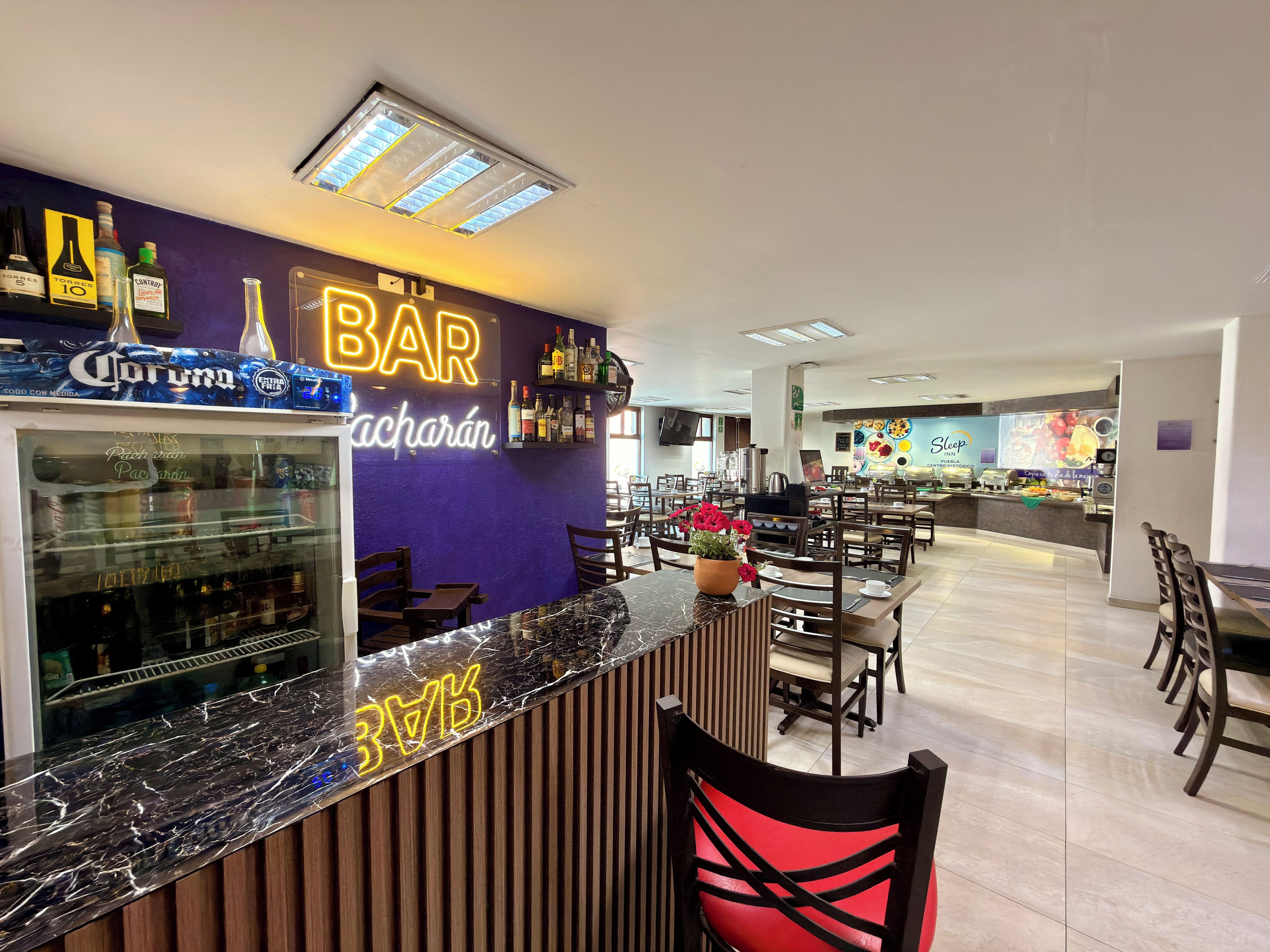 Una zona de bar con un letrero de neón, un refrigerador lleno de bebidas y un área de estar con mesas y sillas.
