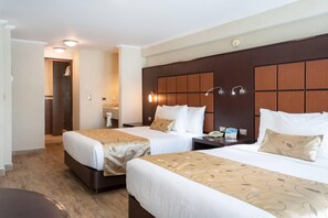 Habitación de hotel con dos camas, cabecera de madera, lámparas de noche, mesitas de noche y un baño visible a través de una puerta abierta.