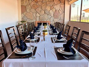 Una larga mesa de comedor preparada para una comida, con sillas a ambos lados, un muro de piedra al fondo y una vista a través de la ventana.