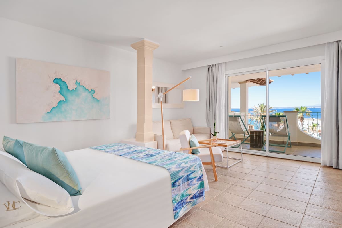 A hotel room with a bed, a desk, a chair, and a balcony with a view of the sea.