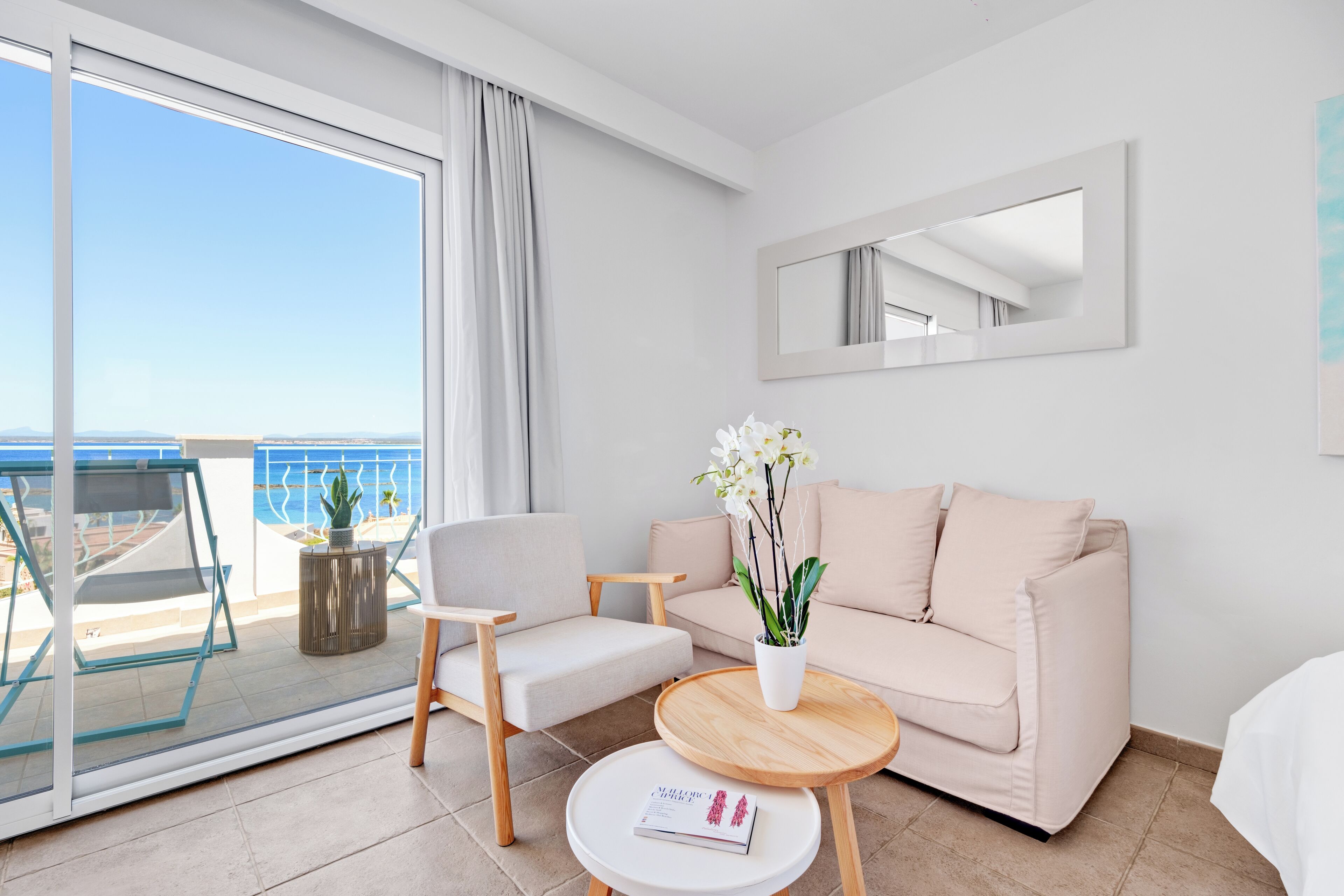 A modern hotel room with a balcony, a sofa, a chair, a small table, and a potted plant.