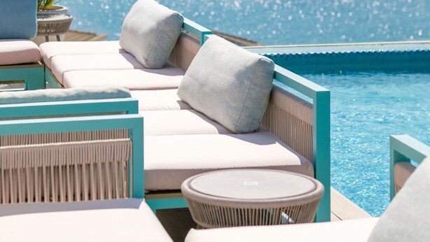 Zona junto a la piscina con sillones de descanso y una mesa, con vistas al mar azul y un velero a lo lejos.