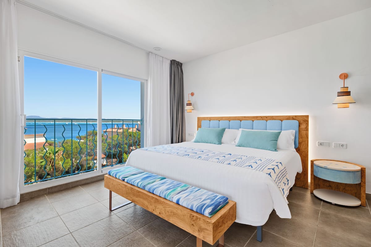 A bedroom with a large bed, a bench, a bedside table, and a view of the sea.