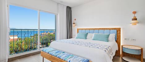 A bedroom with a large bed, a bench, a bedside table, and a view of the sea.