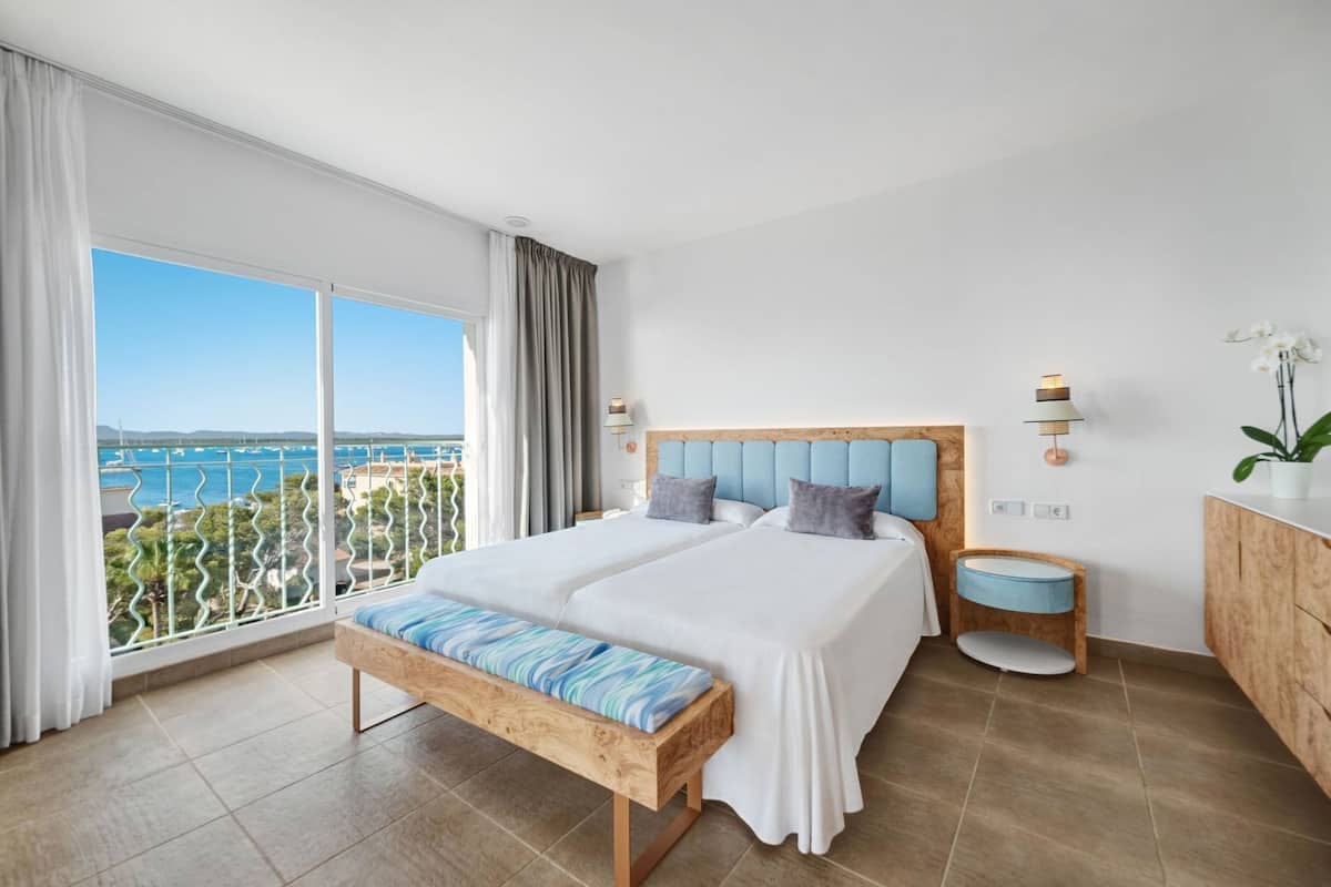 A bedroom with two beds, a bench, a nightstand, a lamp, and a view of the sea.
