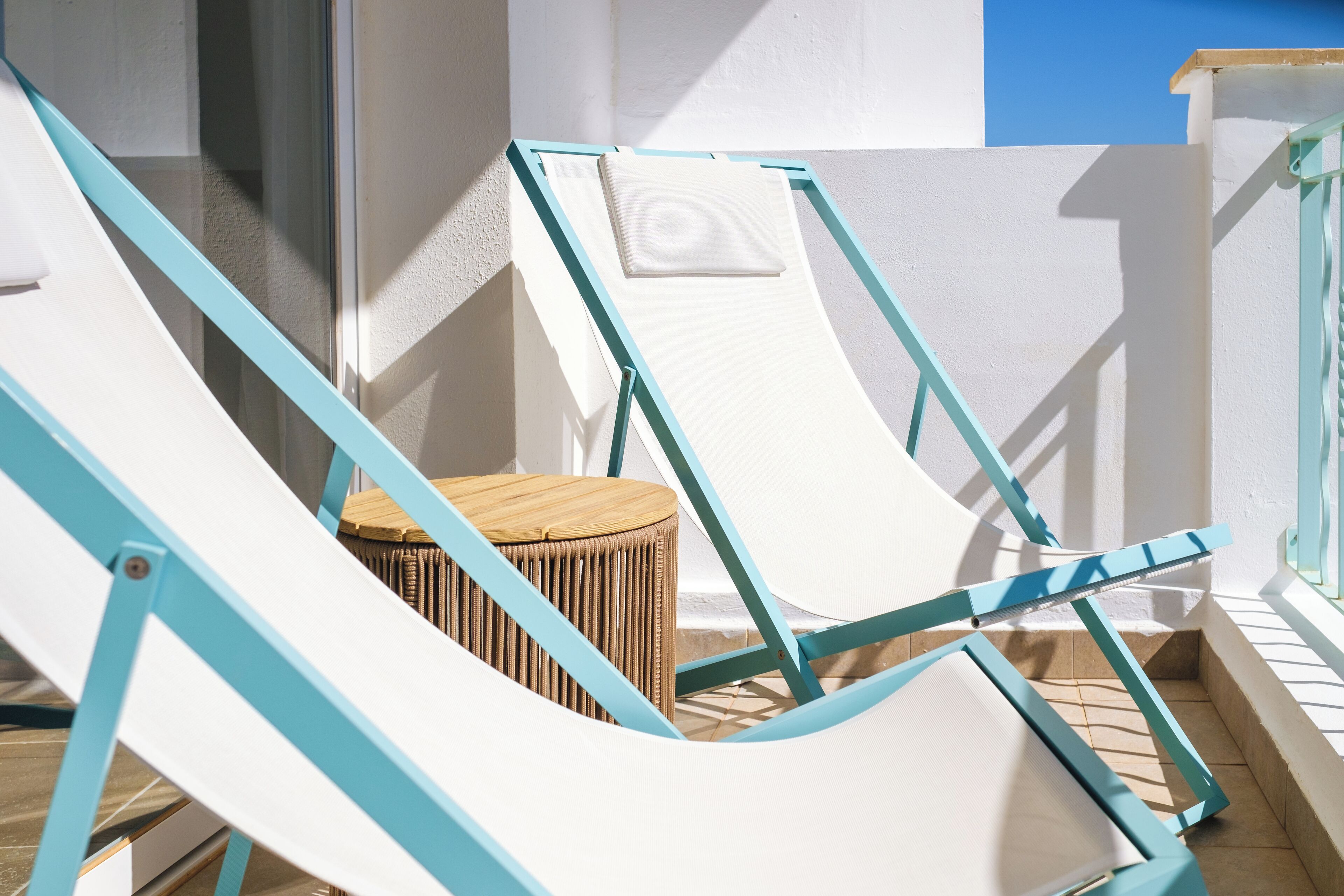 Un balcon ensoleillé avec deux chaises longues blanches et un petit tabouret en bois.