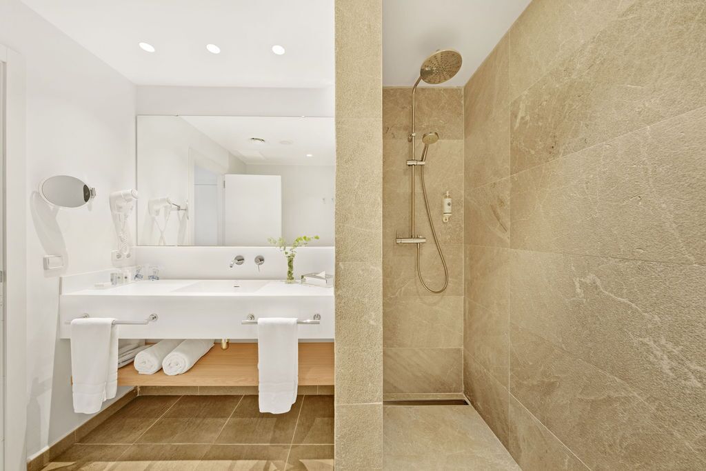 Un baño moderno con un espejo grande, lavamanos blanco y ducha con baldosas color beige.