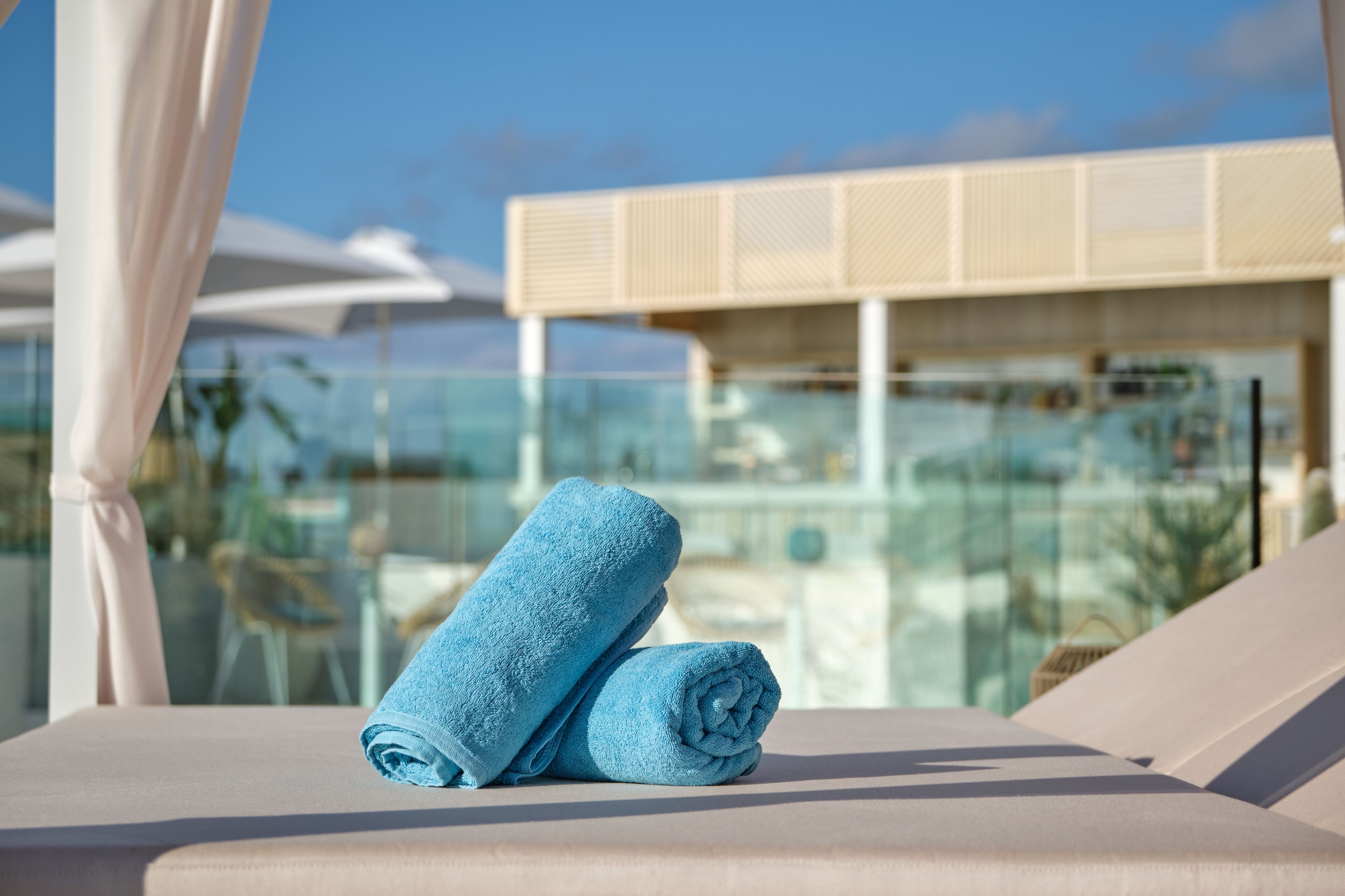 Un transat avec une serviette bleue roulée, une balustrade en verre et un bâtiment moderne en arrière-plan.