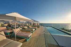Terraza soleada con sillones reclinables y sombrillas, con vistas a una piscina y el océano.