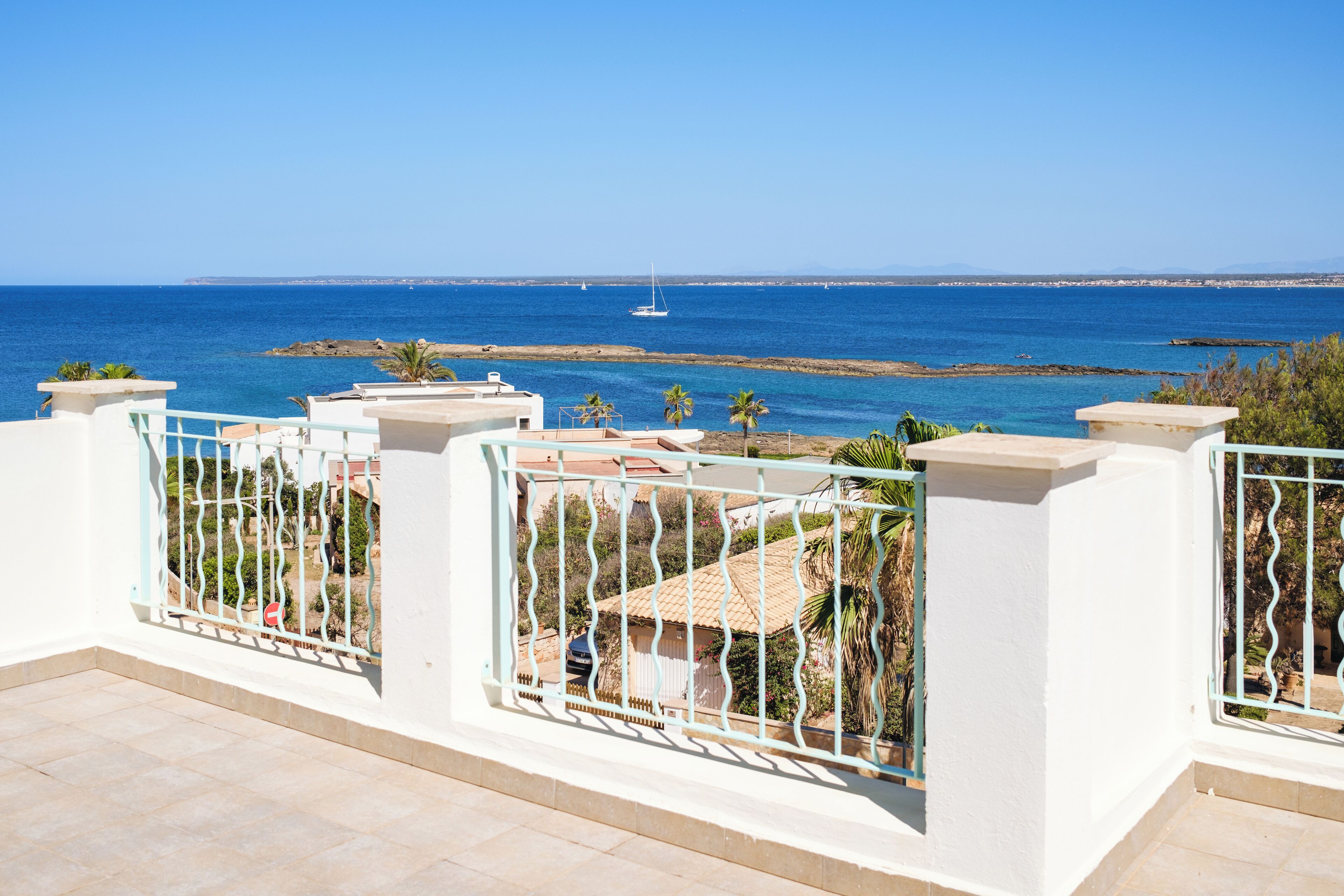 A balcony with a view of the sea, a sailboat, and a distant shoreline.