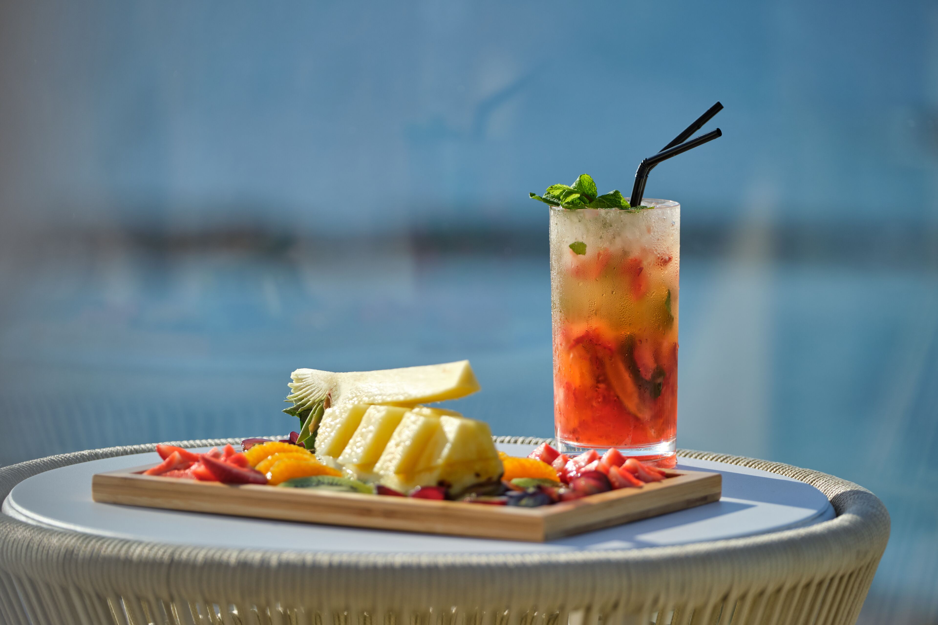 A fruit platter and a glass of iced drink on a wicker table by a pool.